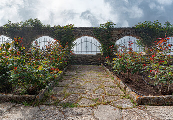 Naklejka premium A paved stone path leads toward a scenic stone wall with arches, wrought iron fencing, and blooming rose bushes under a cloudy sky.