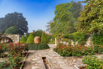 Naklejka premium A beautiful classic European garden features stone pathways, lush green trees, decorative bushes, and a terracotta urn centerpiece under a clear blue sky.
