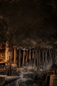 Blacksmith hammers, tongs hanging on rack at old soot covered stone wall of dark rustic forge with anvil horn