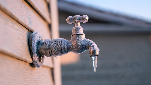 Frozen outdoor spigot stands encrusted with heavy frost during severe cold weather. Small icicle hangs from metal valve nozzle, highlighting plumbing risk on residential house wall.
