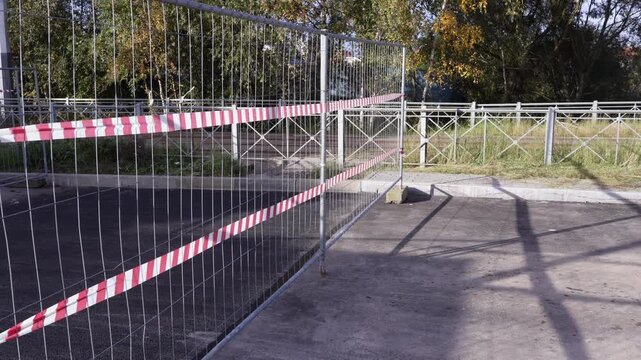 Metal mesh fence with diagonal red-and-white hazard tape cordons off an empty paved area amid greenery and trees