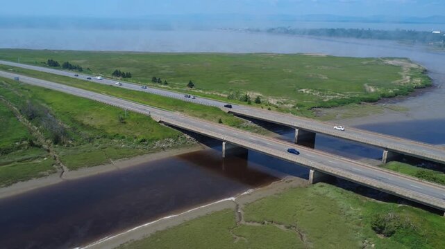 Aerial drone footage shows smooth-flowing traffic crossing two parallel road bridges over the Rivi&egrave;re-du-Loup mouth at low tide amid morning mist in Quebec, Canada.