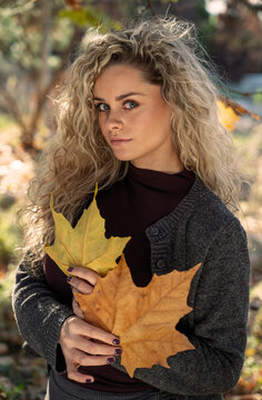 A young woman with curly blonde hair holds two large autumn leaves while looking directly at the camera.