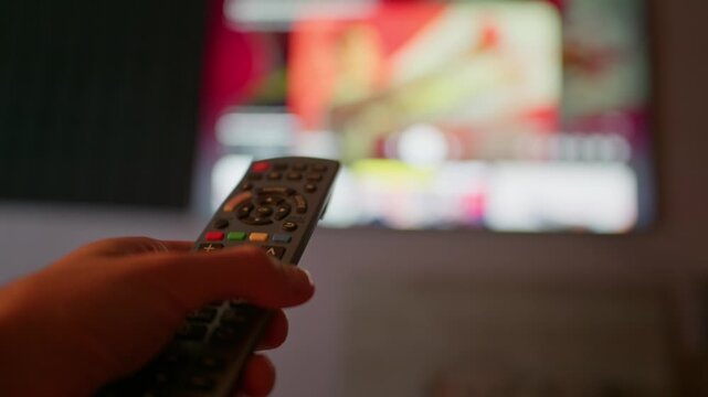 A person operates a television remote control in a living room close-up. Shallow depth of field highlights home entertainment and streaming media concepts for lifestyle projects.