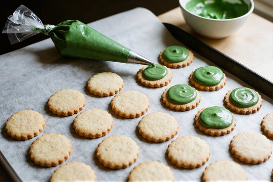Baking sheet with round shortbread cookies topped with green icing, piping bag and bowl of frosting on parchment paper, natural holiday cookie decorating close-up