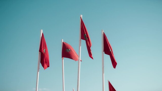 Red Moroccan flags with green pentagrams flutter in the wind on white flagpoles against a clear blue sky.