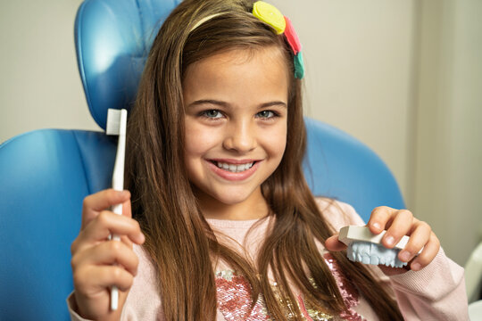 A smiling young girl sits in a dental chair while holding a toothbrush and a dental model clone of teeth. Learn how to brush teeth properly 