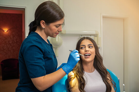 A female dentist uses a dental mirror to closely examine the braces of a smiling young patient.