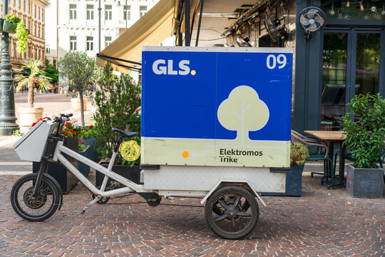 Budapest, Hungary - October 9, 2026: GLS delivery. An electric cargo tricycle with a large blue delivery box is parked on a cobblestone street beside an outdoor cafe in Budapest