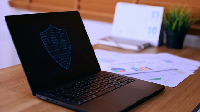 Laptop computer displaying cyber security shield icon with binary code on screen at wooden office desk. Professional data protection and network safety technology concept background.