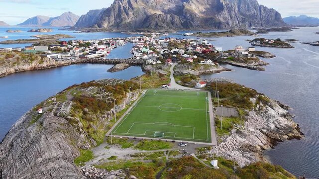 Aerial view of Henningsvaer, a picturesque fishing village in Lofoten, Norway. Colorful houses dot the coastline, surrounded by majestic mountains and the iconic football field