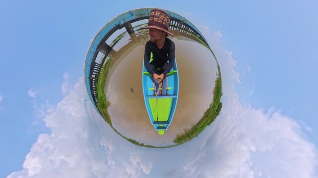 Thale Noi Lake Chaloem Phrakiat Bridge Man in a Patterned Hat Rides a Blue and Green Wooden Boat on a Muddy River Near a Bridge, Spherical Little Planet View With Clouds and Green Vegetation for Outdo
