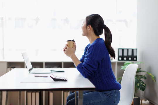 Asian woman taking a refreshing coffee break from work, sitting at her home office desk, relaxing and contemplating during a busy remote workday