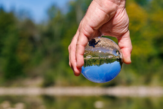 Hand holding a clear crystal lens ball reflecting an inverted lake landscape with forest, rocks and blue sky, blurred green autumn nature background, environment concept