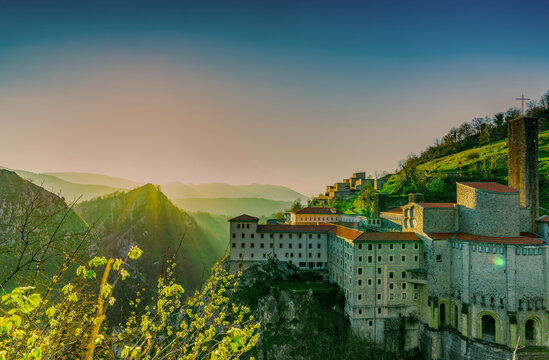 Stone buildings on a rocky mountainside in Spain at sunrise during spring, red tiled roofs and a church tower above a misty valley, green hills and distant mountain ridges beneath a clear morning sky