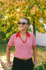 Fototapeta premium Happy young African woman with braided hair and trendy sunglasses smiling outdoors in autumn, wearing a red striped shirt against colorful fall foliage