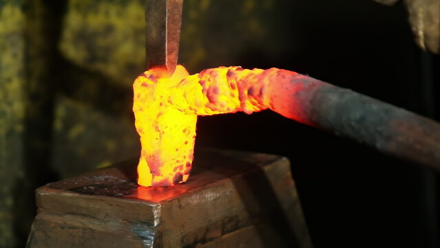 Skilled smith holds incandescent steel billet with metal tongs. Worker carefully aligns glowing material under striking head of powerful hammer