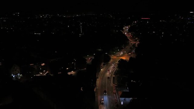 Cars travel curving illuminated road through dark American city at night, orange streetlights tracing winding hillside route above scattered neighborhood lights below. Aerial drone wide shot