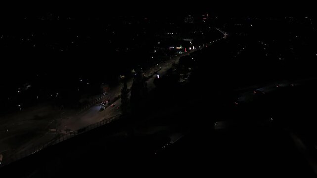 Scattered headlights trace winding overpass through near-total darkness, distant illuminated storefronts and highway bridge glowing above quiet wide American suburb below. Aerial drone wide shot