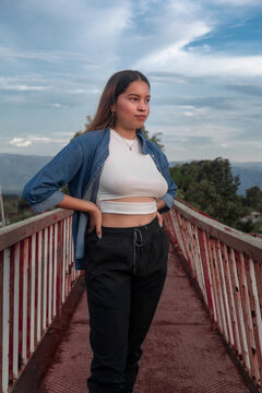 Young Latina woman wearing denim jacket and white crop top posing on footbridge.