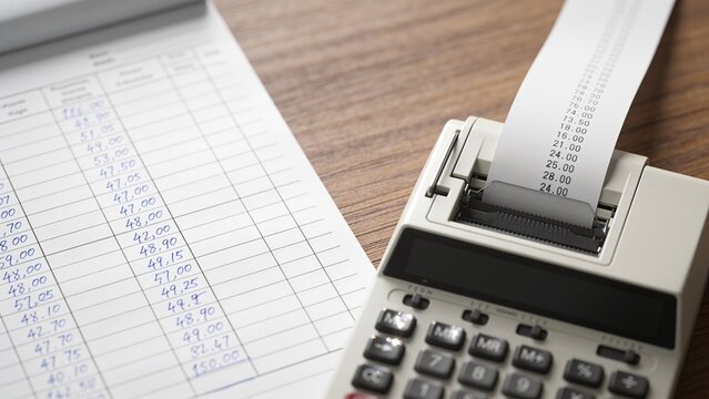Displaying printing adding machine feeding receipt tape on office wood desk, with ledger