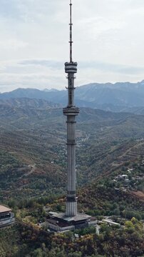 Vertical 9:16 4K drone video close up of iconic television tower on Kok Tobe hill in Almaty, Kazakhstan at sunny September morning, revealing colorful red and yellow autumn trees and Tian Shan mount