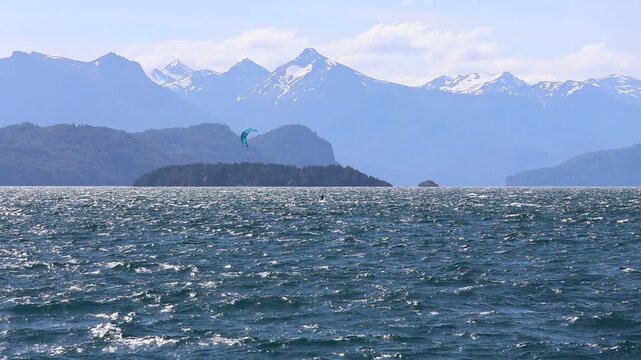 A person windsurfing on Lake Nahuel Huapi on a sunny summer day, contrasting with the snow-capped mountains in the background.