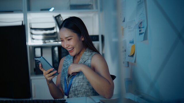 A woman laughing at a smartphone during a mobile office break gestures near her chest with her free hand, keeping the smartphone nearby during an everyday personal leisure moment at the desk indoors.