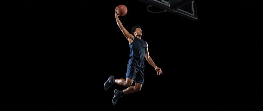 Young basketball player in a dynamic action pose with a basketball, photographed from a ground-level perspective against a solid black background, illustrating athleticism and movement, with copy spac