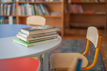 Stack of children's books on round table and small wooden chair in bright school or library, bookshelves in background. Early childhood education, back-to-school, reading time, learning environment. © Natallia