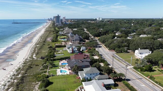 Flight along a coastal road passes oceanfront houses with pools toward a house under construction with an excavator, beach and pier beyond in Myrtle Beach, South Carolina. Aerial approaching shot