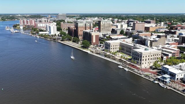 A lone sailboat drifts past the Alton Lennon Federal Building and its palm lined riverwalk lawn in Wilmington, North Carolina, brick downtown blocks rising behind. Aerial view
