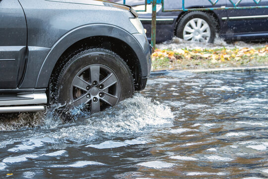 A car navigates a flooded city street as rain pours down, creating puddles and causing difficulties for vehicles in the area