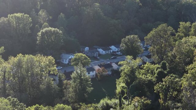 Small ranch houses line a street in a wooded valley in Lynchburg, Virginia, softened by hazy backlit sun and framed by leafy trees in early autumn, compressed by a long lens. Aerial view