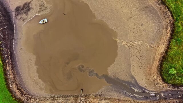 Aerial timelapse of a small boat stranded in a drying muddy pond, surrounded by grass