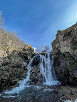 Mountain waterfall cascading into the rocky pool in rural Morocco, bare trees and rugged cliffs, blue sky