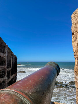 Rusty coastal cannon framed by stone ramparts, overlooking Atlantic ocean waves in Essaouira, Morocco, blue sky