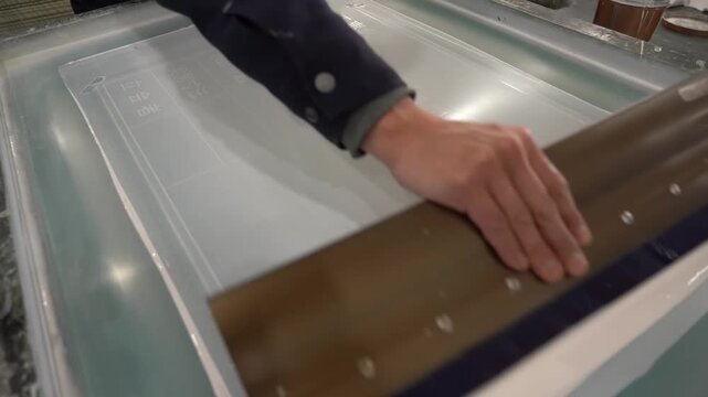 Craftsman pulling a squeegee steadily across a large silkscreen frame to press white ink through the mesh