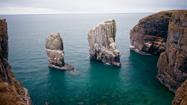 Aerial drone view of towering sea stacks rising from turquoise ocean water between rugged coastal cliffs with layered rock faces and calm open sea near Falmouth Cornwall England UK.