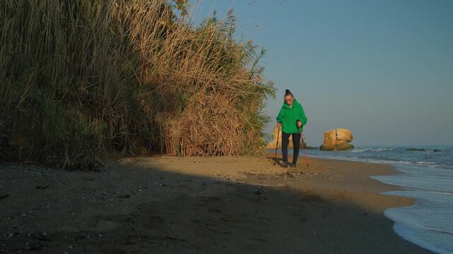 Senior Woman Walks Along Seaside Beach With Trekking Poles. Scandinavian Walking For Eldery People. 