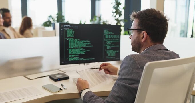 Programmer developer man coding on computer workstation. He reviews documents with a pen as code fills the monitor, aligning software to goals. Professional technology office workflow concept.