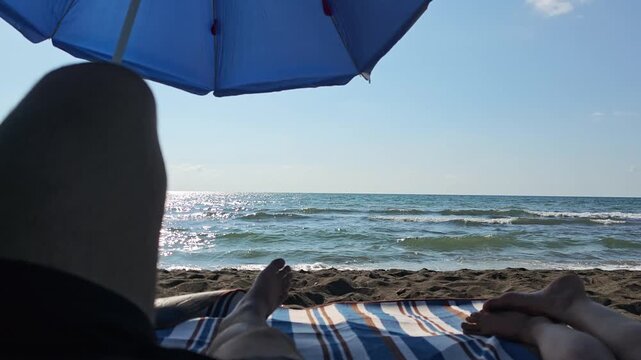 Personal perspective of a young couple relaxing on a sandy beach under a blue umbrella, enjoying their summer vacation together while sunbathing and looking at the calm sea waves