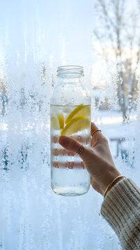 Hand holding glass bottle of lemon detox water against frosty winter window