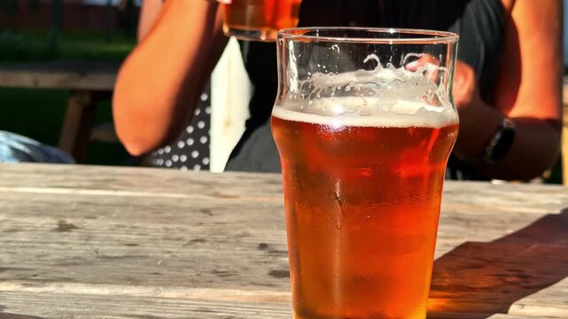 Female hand taking a refreshing glass of cold amber ale from a wooden table on a sunny day