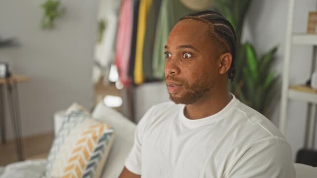 Man biting nails and covering face on sofa in a studio apartment with pillow and clothing rack; anxiety tension.