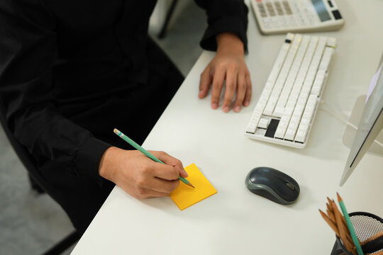 Closeup businessman writing notes on sticky note at office desk with computer, keyboard, and whiteboard in background