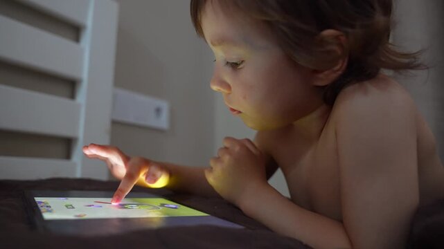 A boy is lying on a bed, intently focused on a tablet screen. The boy's small hand touches the screen, highlighting the glow of the device in a dimly lit room.