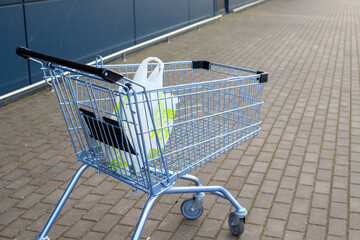 Light blue metal shopping cart carrying single plastic grocery bag stands on paved ground outside supermarket wall. Retail trade, consumerism, purchasing power, and grocery store service © yaviki