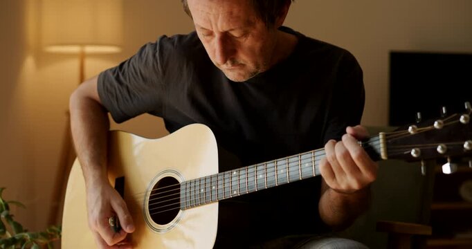 Man playing an acoustic guitar at home, close-up of hands strumming and fretting the strings, hobby and music concept in a cozy warm interior. 4K footage.