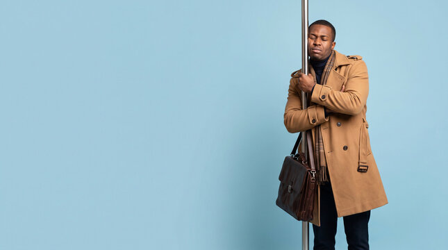 Man leans against a pole, resting his head. He wears a tan coat and carries a brown bag. His expression shows tiredness or calm repose. Simple background emphasizes the subject's quiet moment